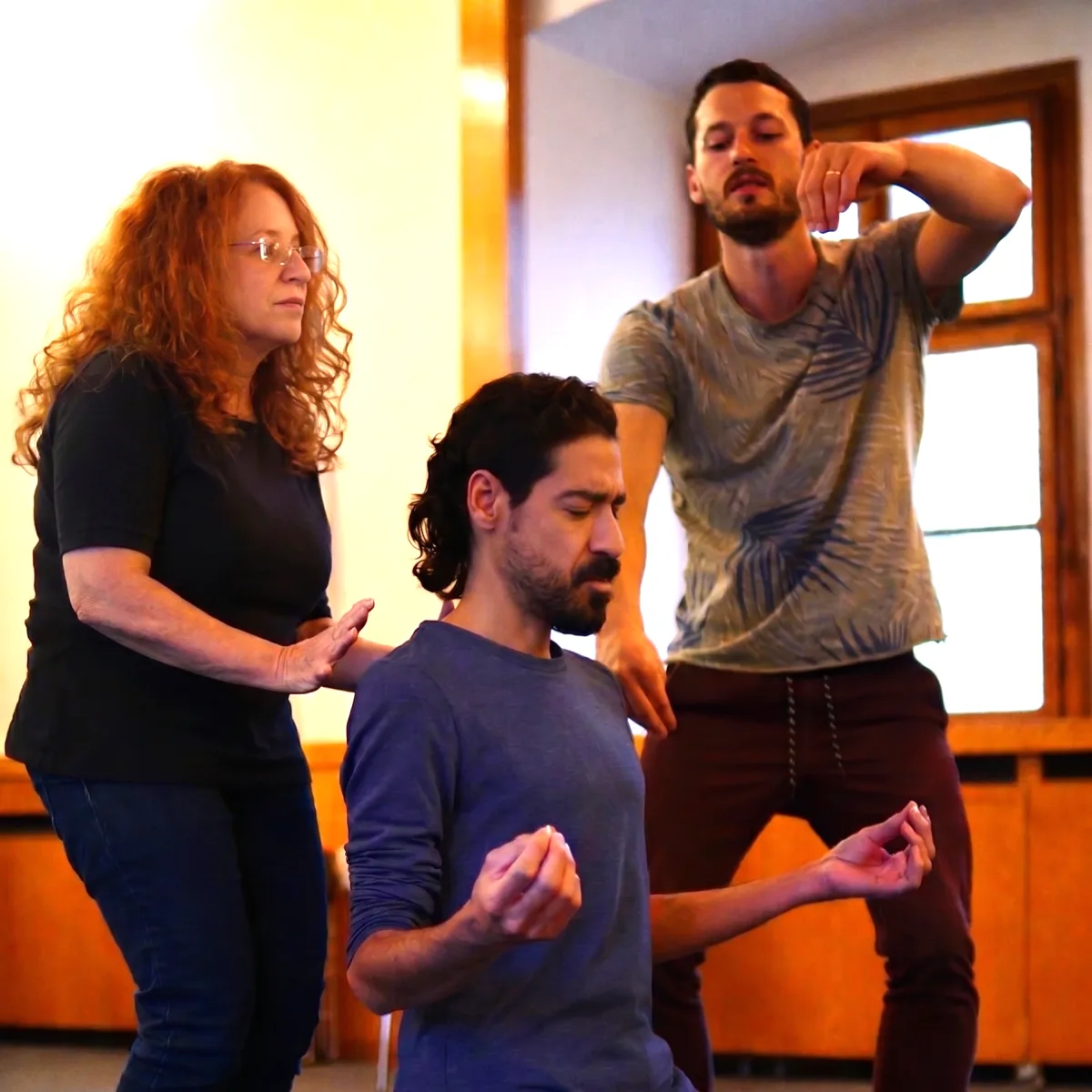 A facilitator guides a participant through a gesture during a Playback Theatre enactment exercise, with another practitioner looking on.