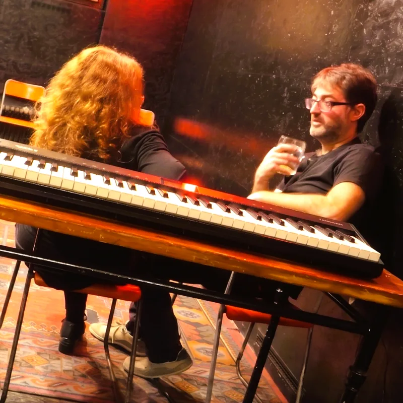 Two people talking beside a keyboard at an evening Playback Theatre venue.