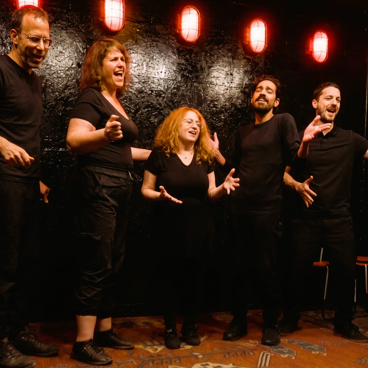 Five Playback Theatre ensemble members singing together on stage, lit by red lantern lighting.